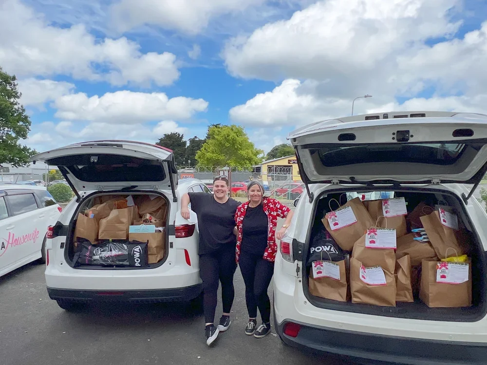 Two women pose with open car trunks filled with paper bags of donations in a parking lot.