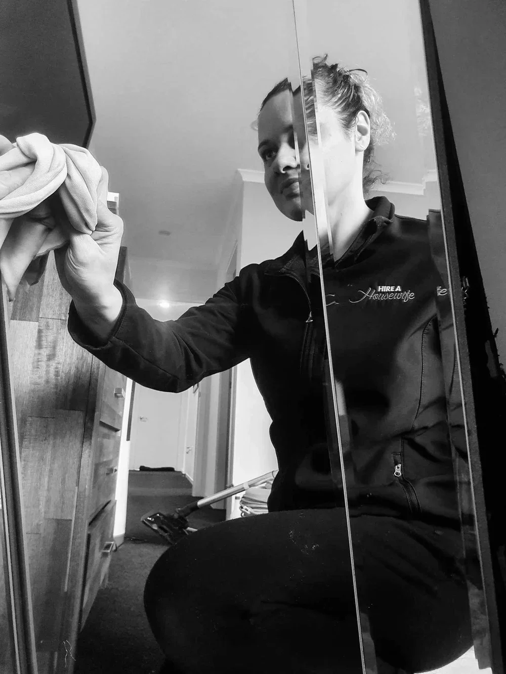 A woman cleaning a mirror with a cloth, taking a polaroid selfie in black and white, wearing a black jacket with the logo 'HIRE A Housewife'.