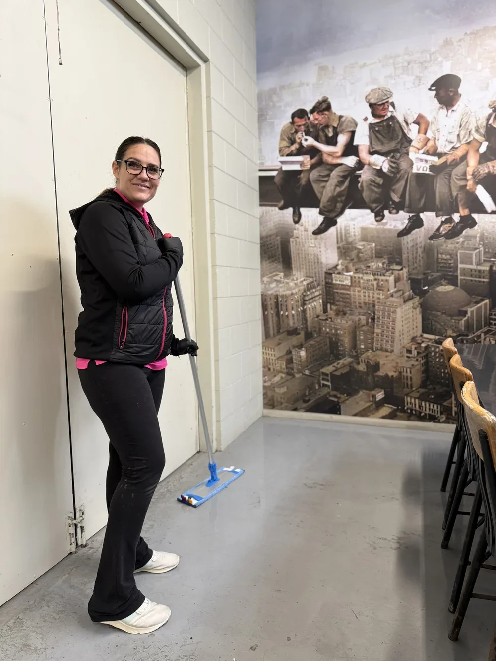 A woman standing indoors next to a wall, holding a mop, smiling at the camera. Behind her is a large mural of people sitting on a ledge high above a cityscape.