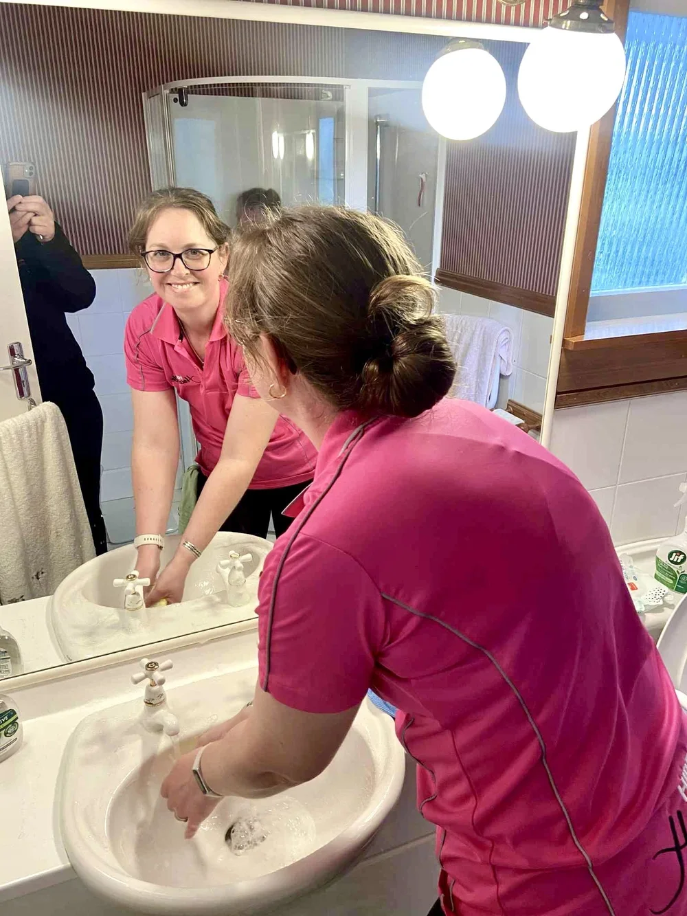 A woman in a pink uniform washing her hands in a sink, smiling while looking at her reflection in the mirror.