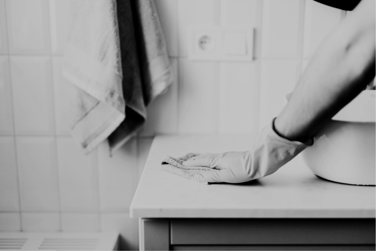 Person wearing a glove and using a cloth to wipe a surface in a bathroom with tiled walls, towels hanging, and a sink.
