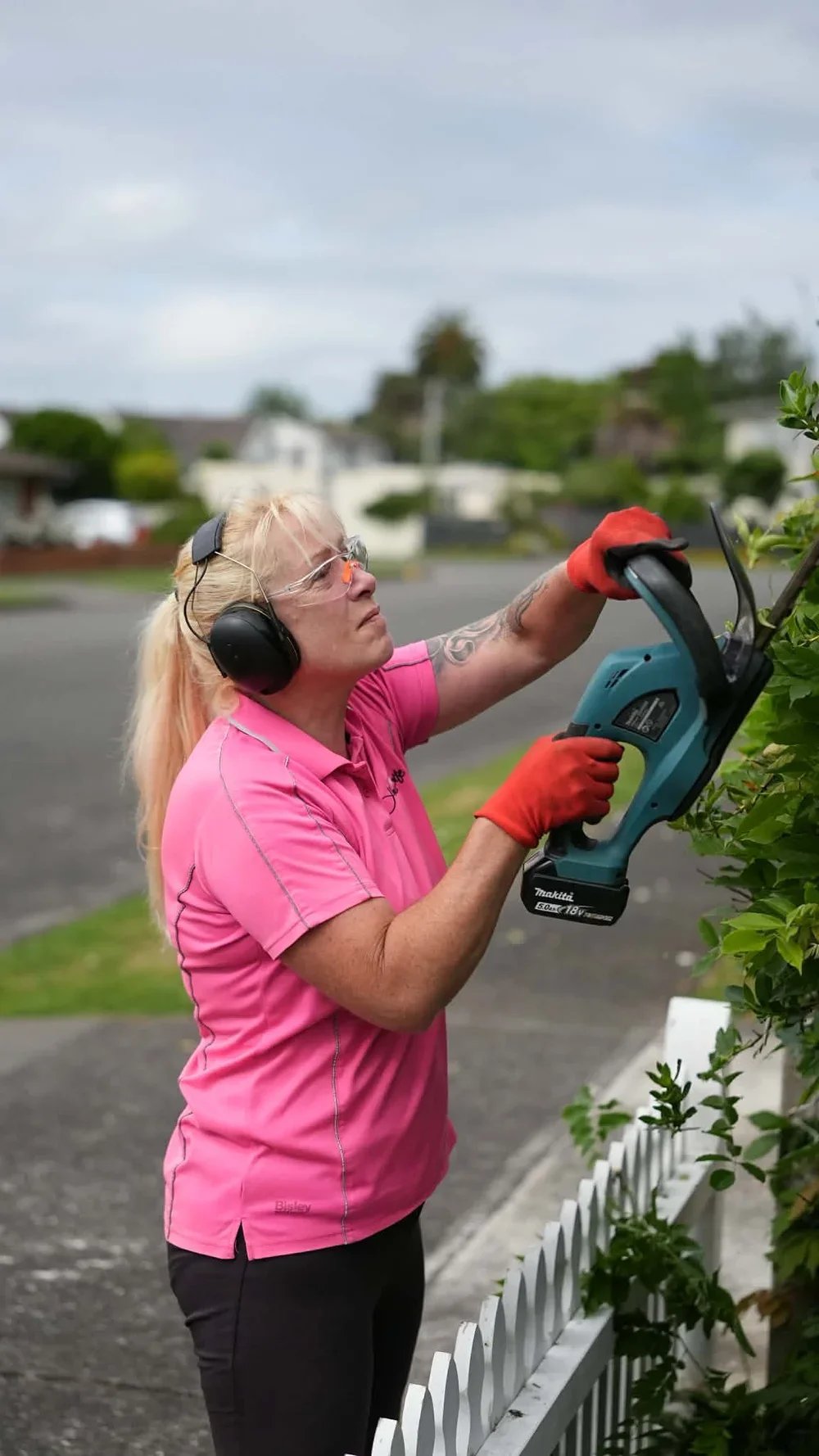 A woman with blonde hair, wearing safety glasses, a pink shirt, orange gloves, and headphones, is trimming a hedge with a power tool outdoors.