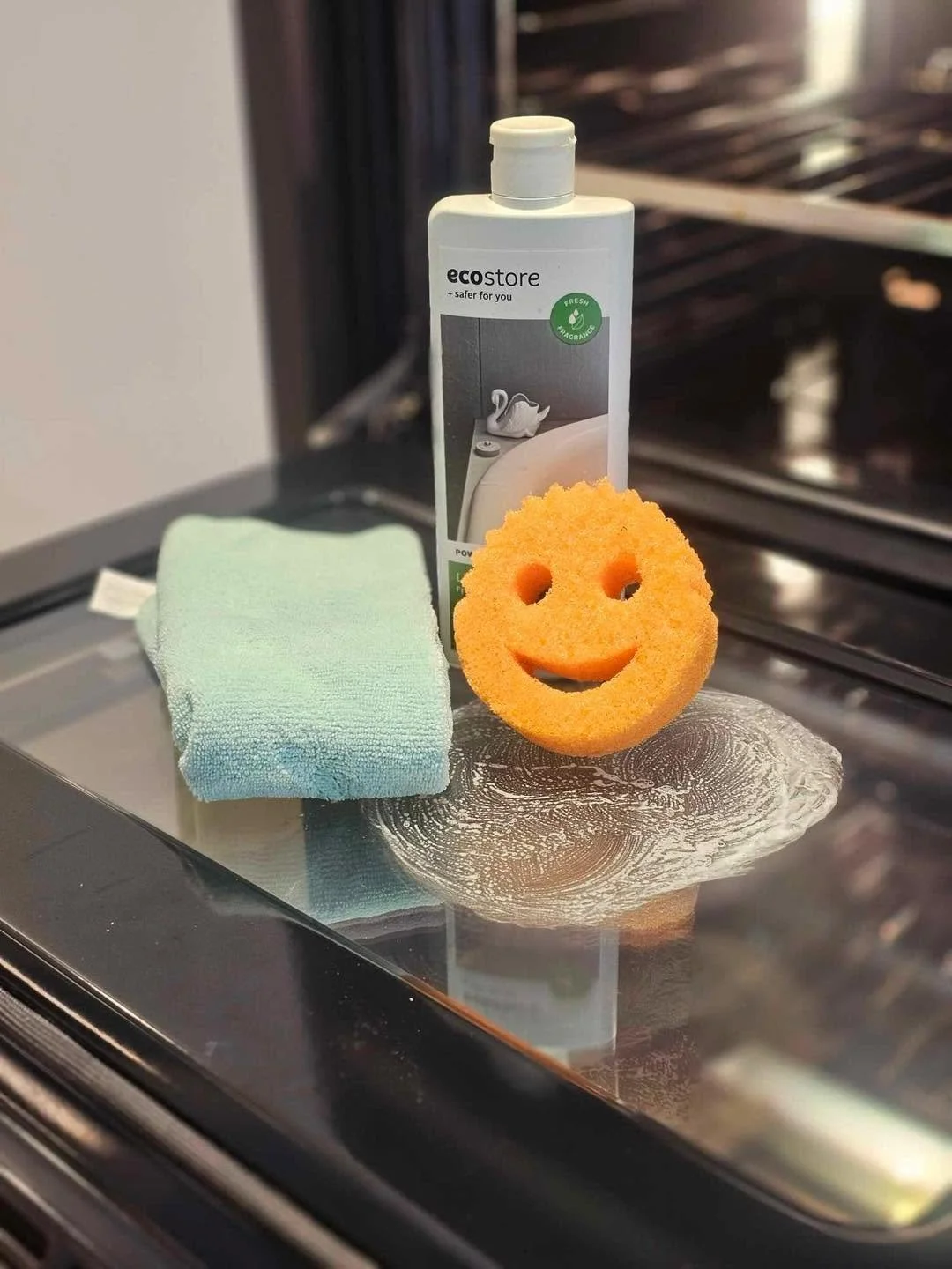 A cleaning sponge with a smiley face, a green cleaning cloth, a bottle of Eco store cleaning product, and a cleaning wipe on a glass stove surface.
