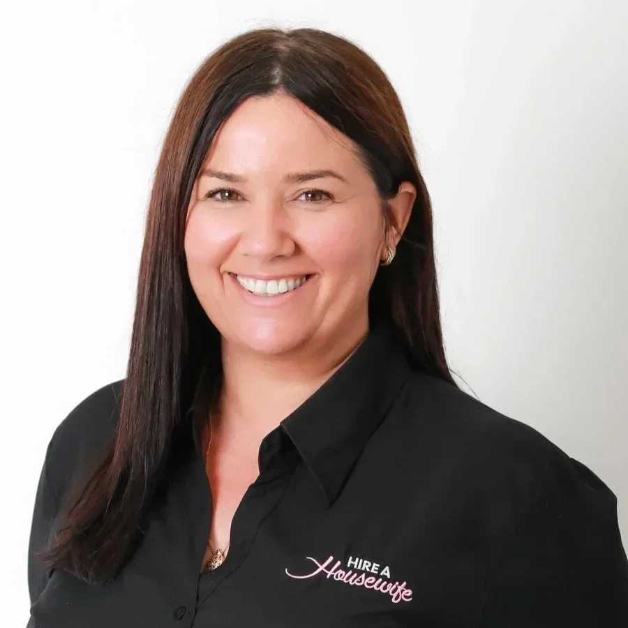 A woman with long dark brown hair, smiling, wearing a black polo shirt with pink and white text that says "HIRE A Housewife".