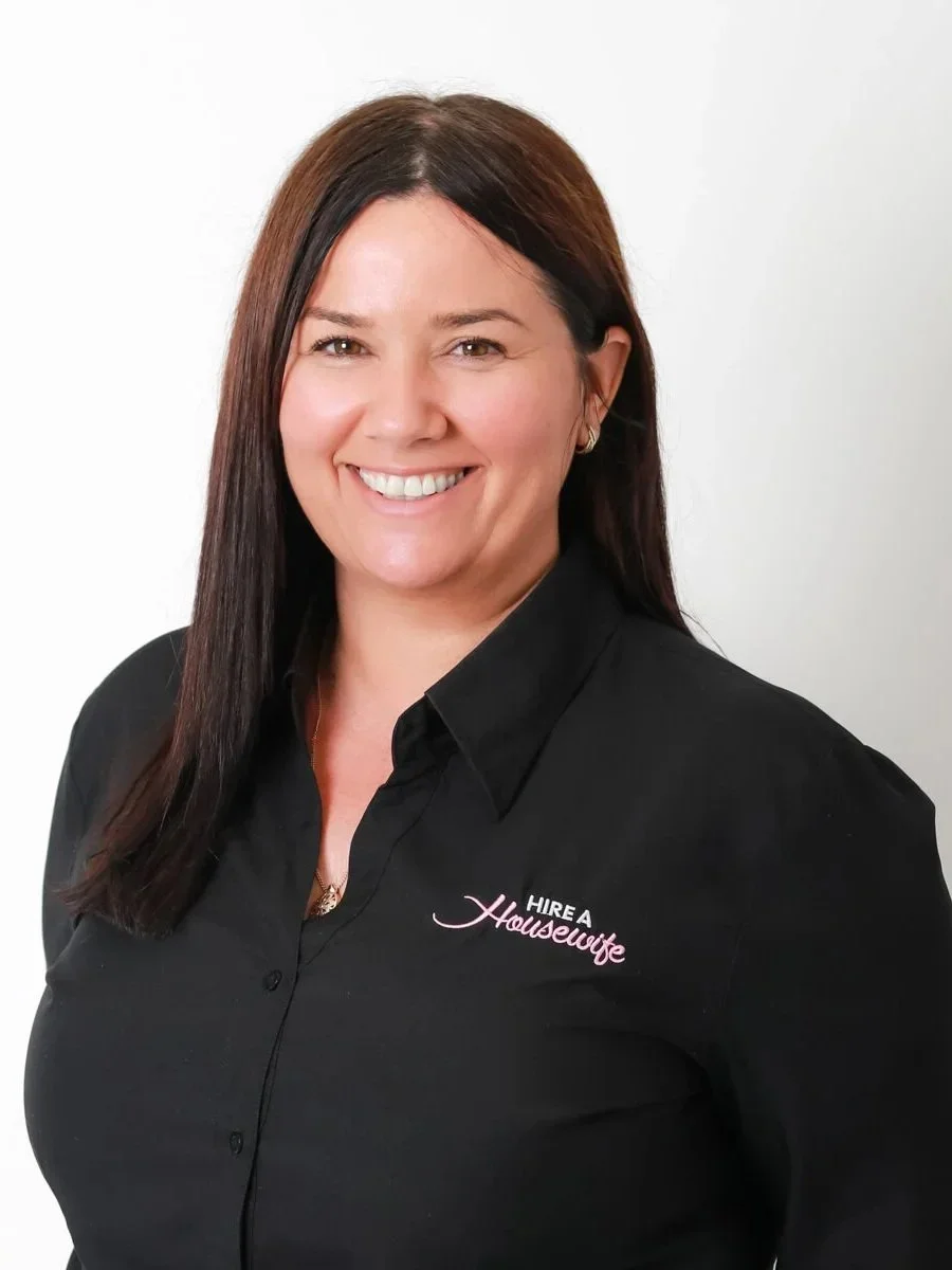 Professional woman with shoulder-length brown hair, smiling, wearing a black collared shirt with the words 'Hire a Housewife' embroidered on the left side, against a plain white background.