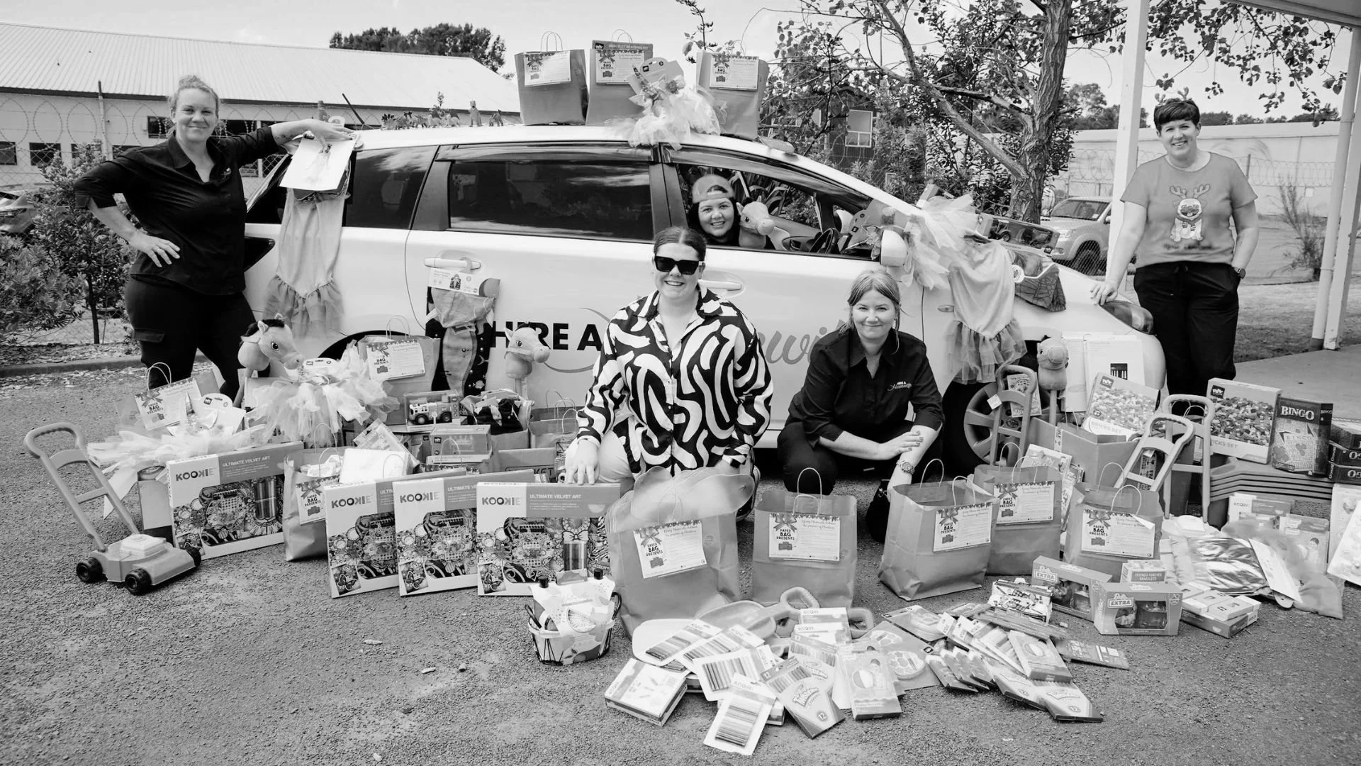 Five women gathering around and in front of a car filled with toys and gift bags, possibly at a charity event or donation drive, with trees and a building in the background.
