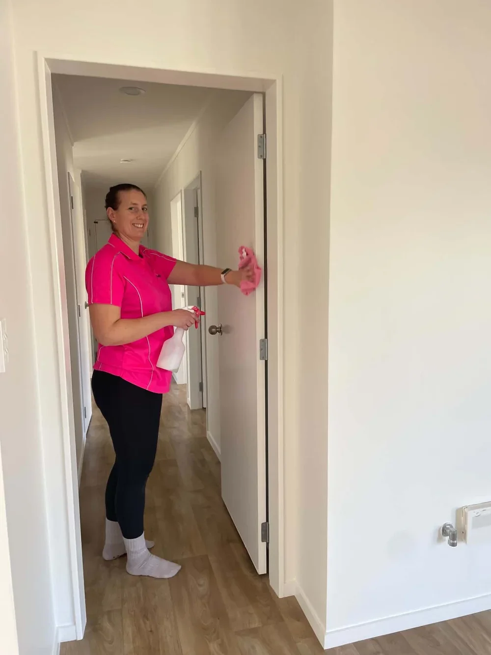 A woman in a pink shirt cleaning a door with a spray bottle and cloth in a hallway of a house.