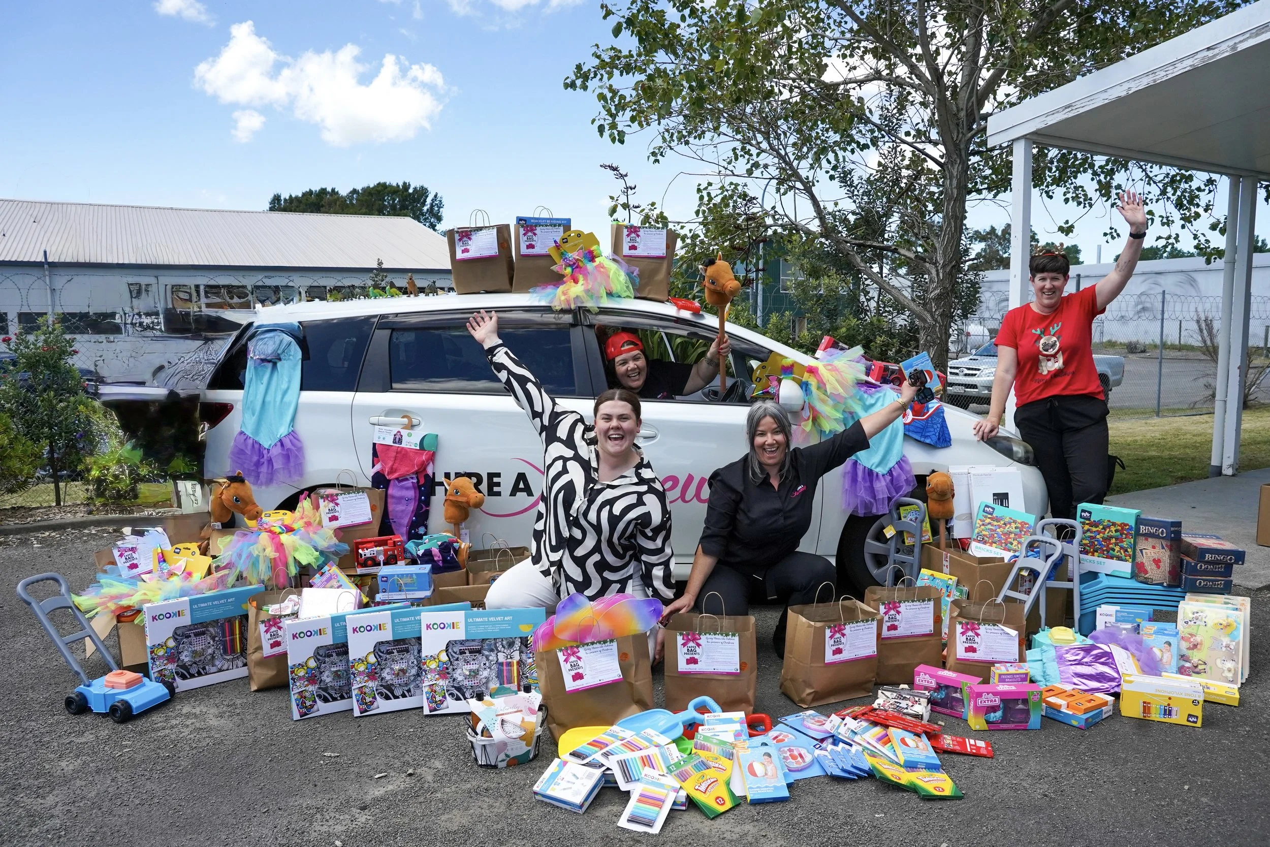Group of people celebrating outside, standing next to a decorated vehicle with toys and gift bags displayed around them during a festive event.