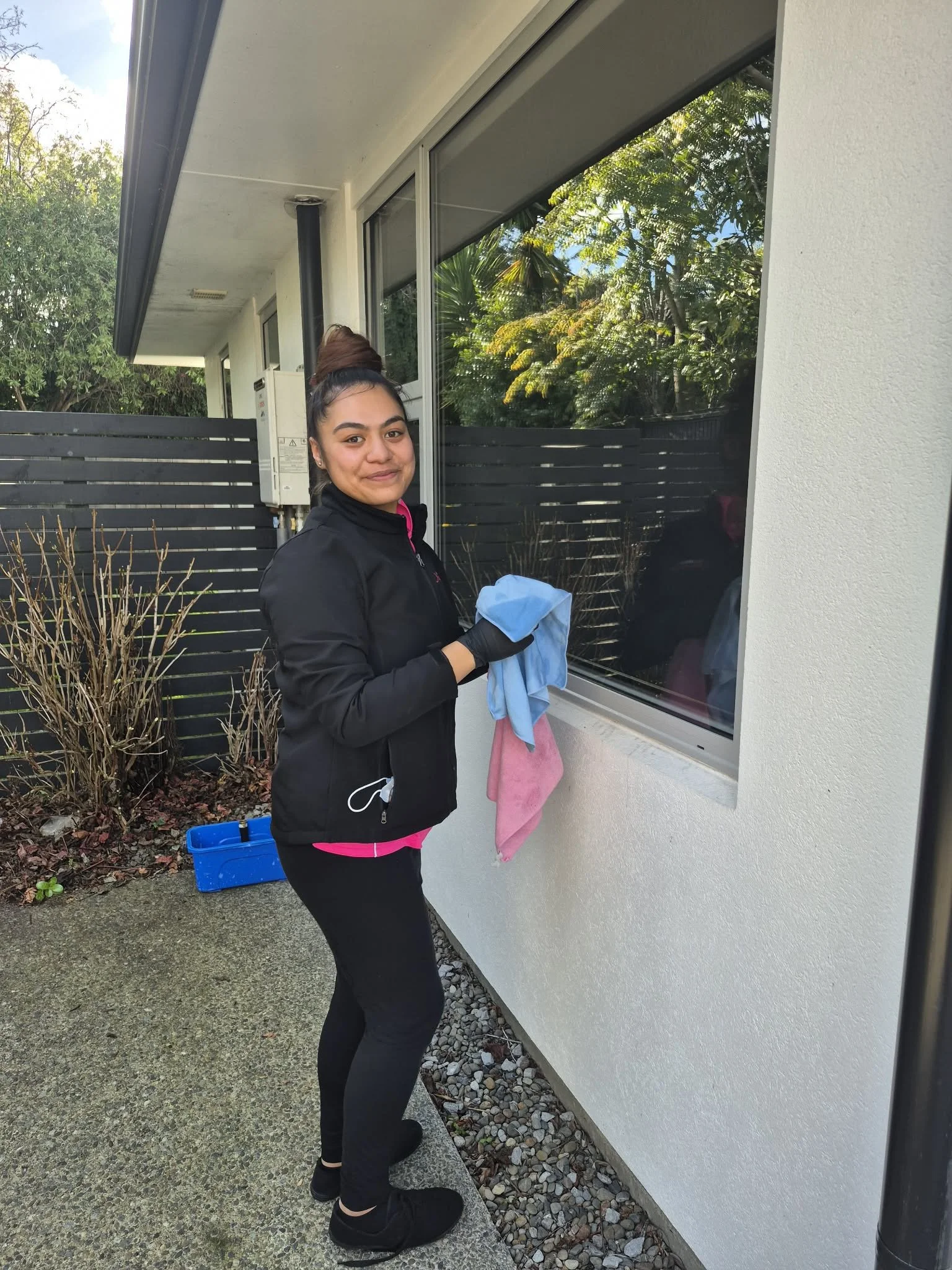 A young woman with dark hair in a bun, wearing a black jacket and black pants, cleaning a large window outside a building on a sunny day. She holds a blue cleaning cloth and a pink towel over her arm. There are bushes, a blue bucket, and a white wall with a black fence behind her.