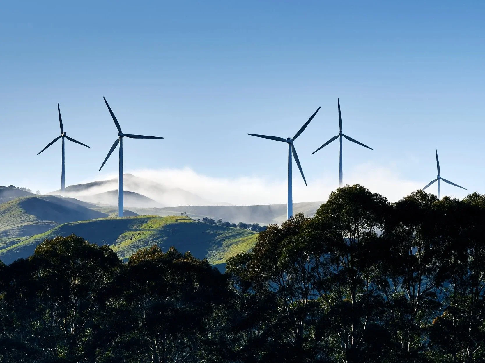 Green hilly landscape with wind turbines in the background under a clear blue sky.