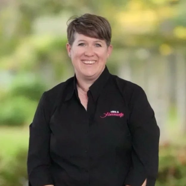 A smiling woman with short brown hair wearing a black collared shirt with pink and white embroidery that says 'Hire a Havanese' standing outdoors with blurred green trees and water in the background.