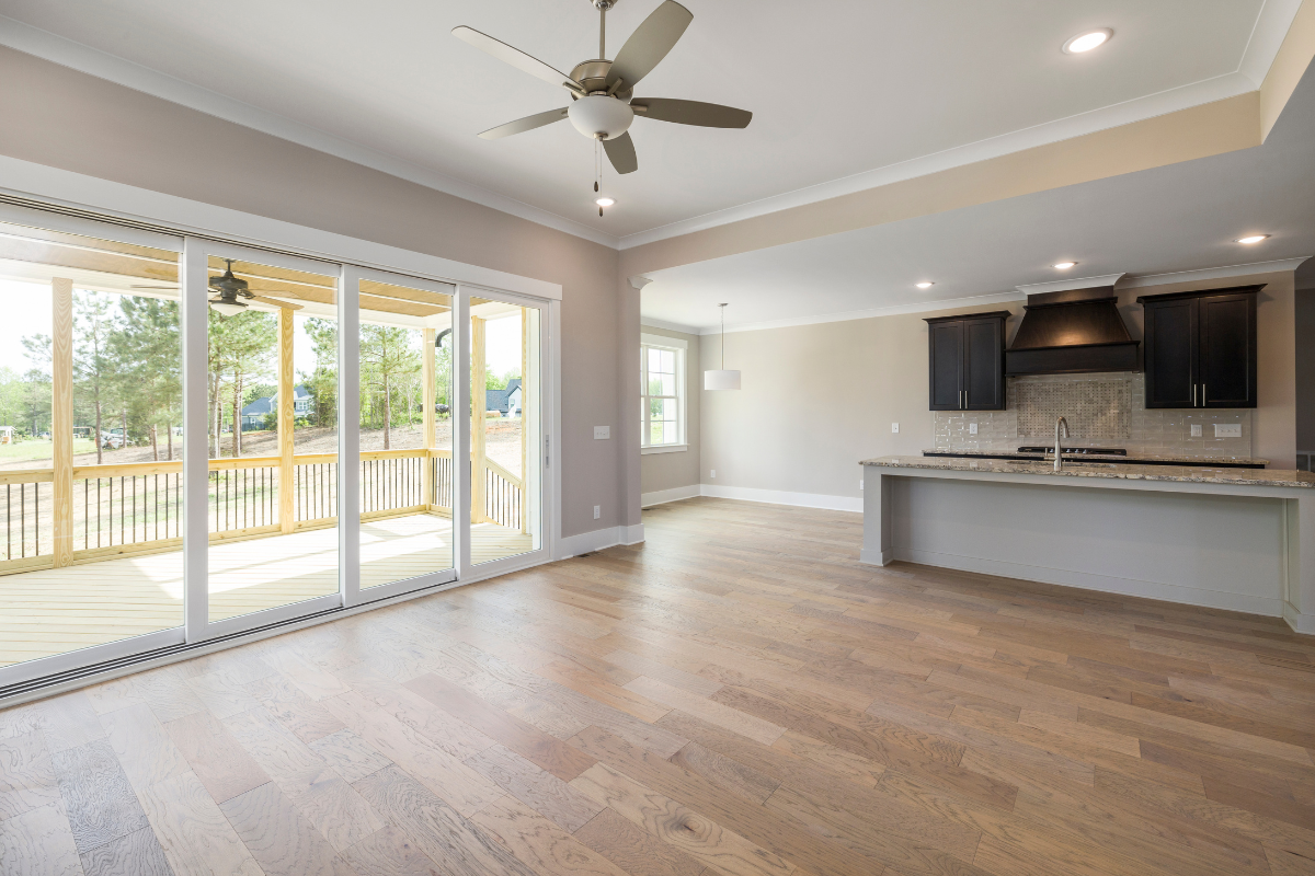 Empty living room with wooden floors, large sliding glass door leading to a deck, ceiling fan, and open kitchen with dark cabinets and granite countertops.