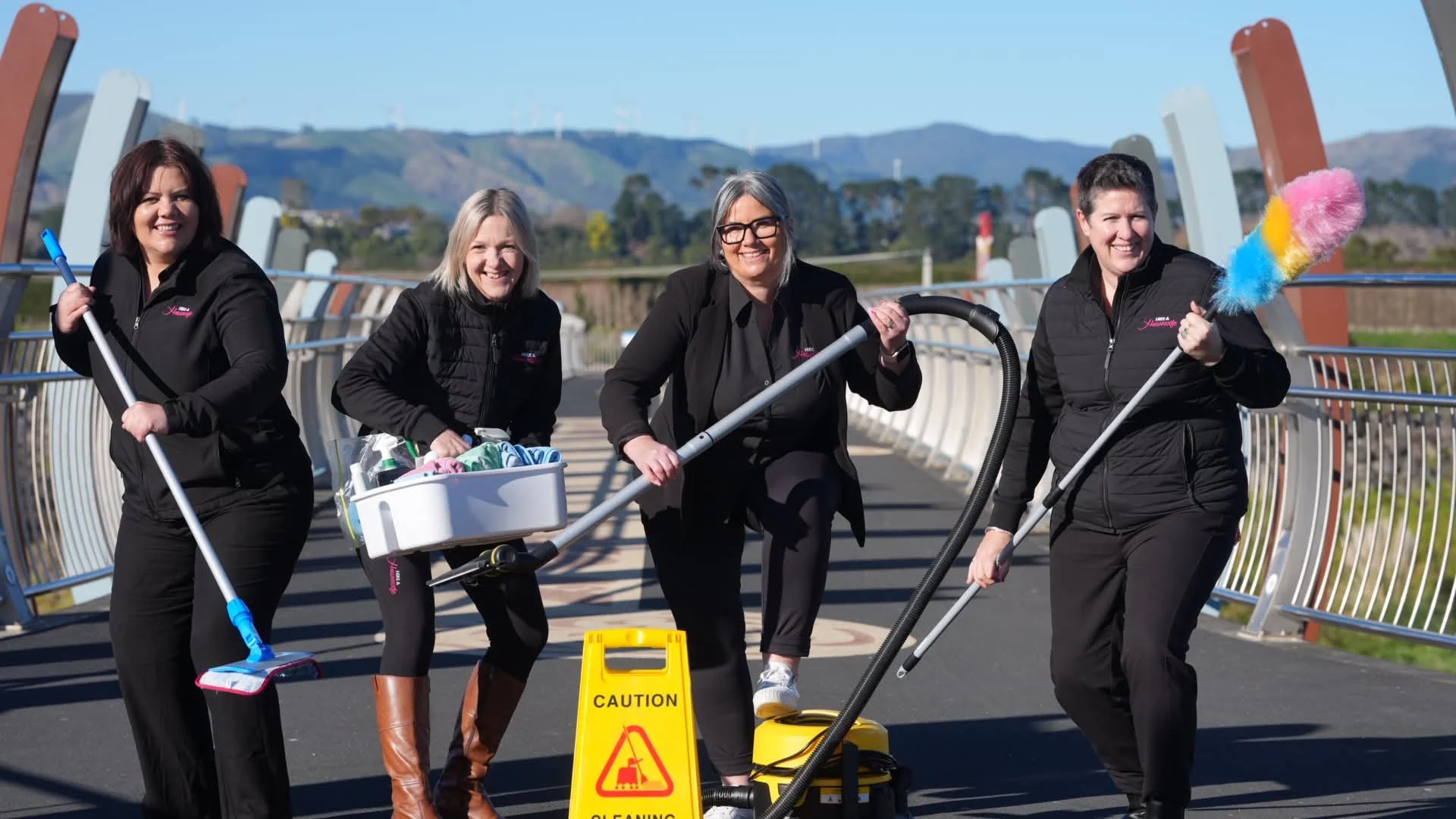 Four women in black jackets cleaning outdoor bridge with mops, vacuum, and cleaning supplies, smiling at the camera, with a yellow caution sign on the ground.