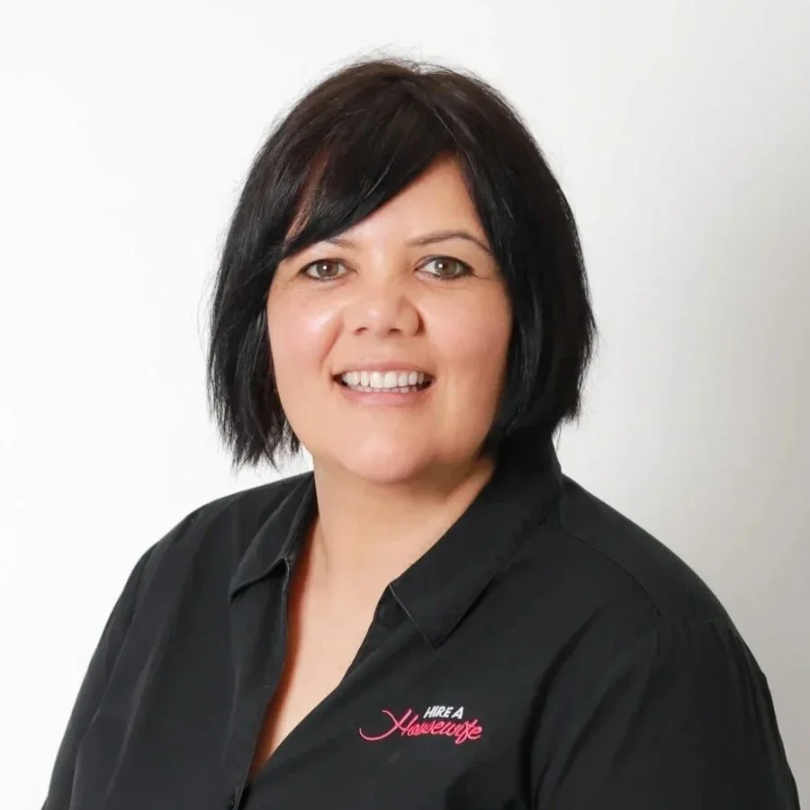 A woman with black hair smiling, wearing a black shirt with a pink logo that says 'Hire a Hairewre'.