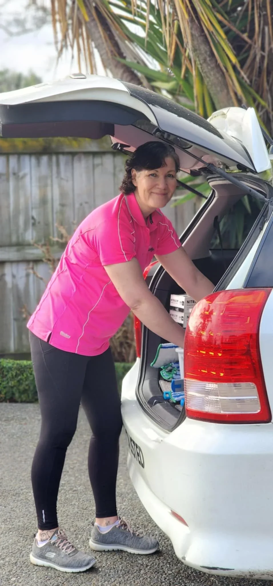 A woman wearing a pink shirt and black pants is loading items into the trunk of a white car, which has its rear hatch open, outdoors with a wooden fence and greenery in the background.