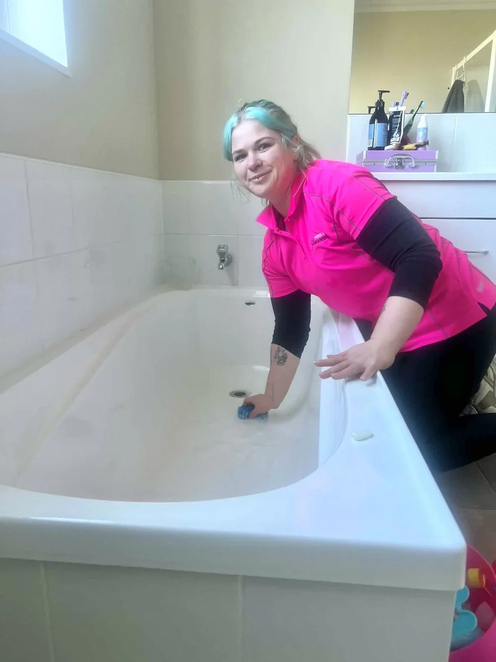 A woman with blue and green hair cleaning a bathtub in a bathroom. She is wearing a pink shirt with black sleeves and has tattoos on her arm. There are bathroom supplies on the counter behind her.
