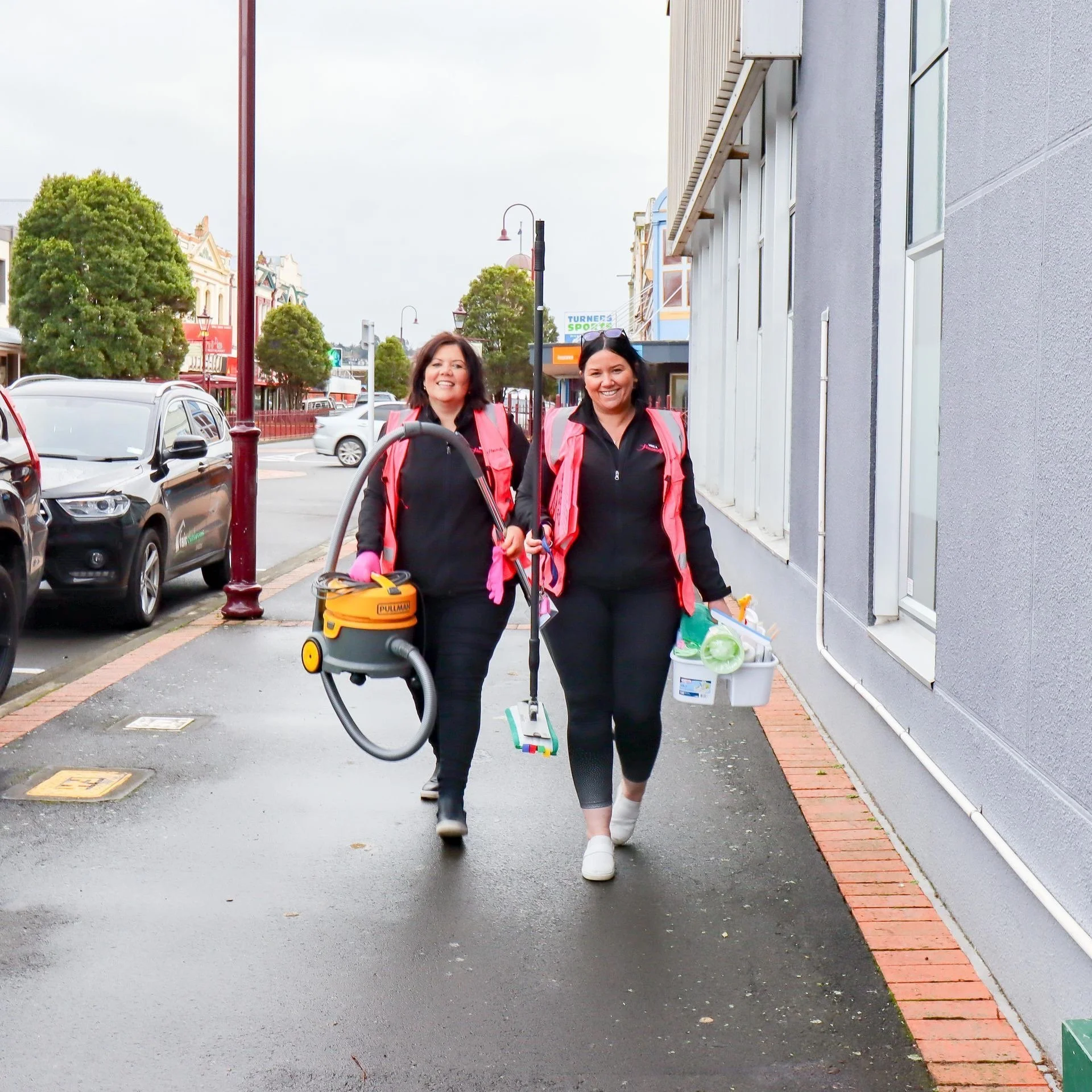 Two smiling women walking on a sidewalk, wearing pink safety vests, with shopping bags and cleaning equipment.