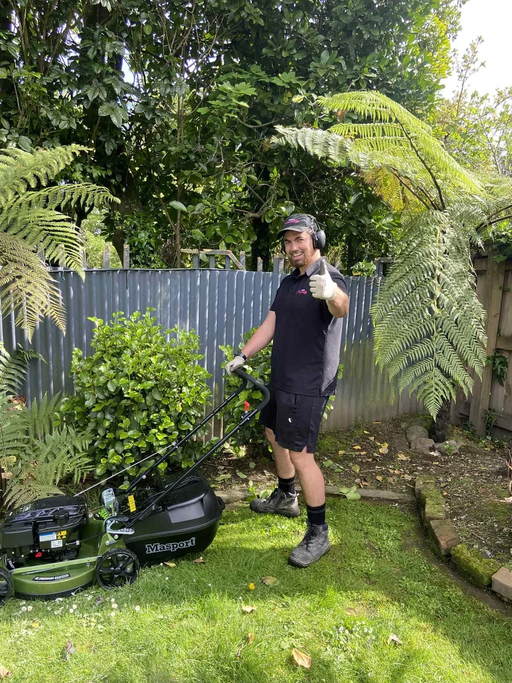 A man wearing a black shirt, black shorts, and work boots, standing on a lawn with a grass trimmer in his hand. He is giving a thumbs-up and smiling at the camera, wearing ear protection and gloves, outdoors in a garden with trees and a metal fence.