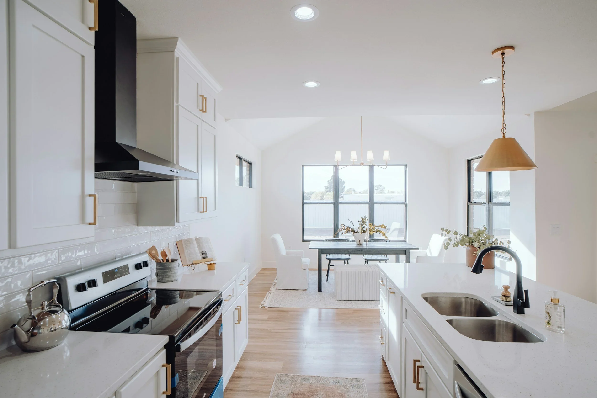 Bright, modern kitchen and dining area with white cabinets, a black stove, a wooden dining table with white chairs, large windows, and a hanging light fixture.