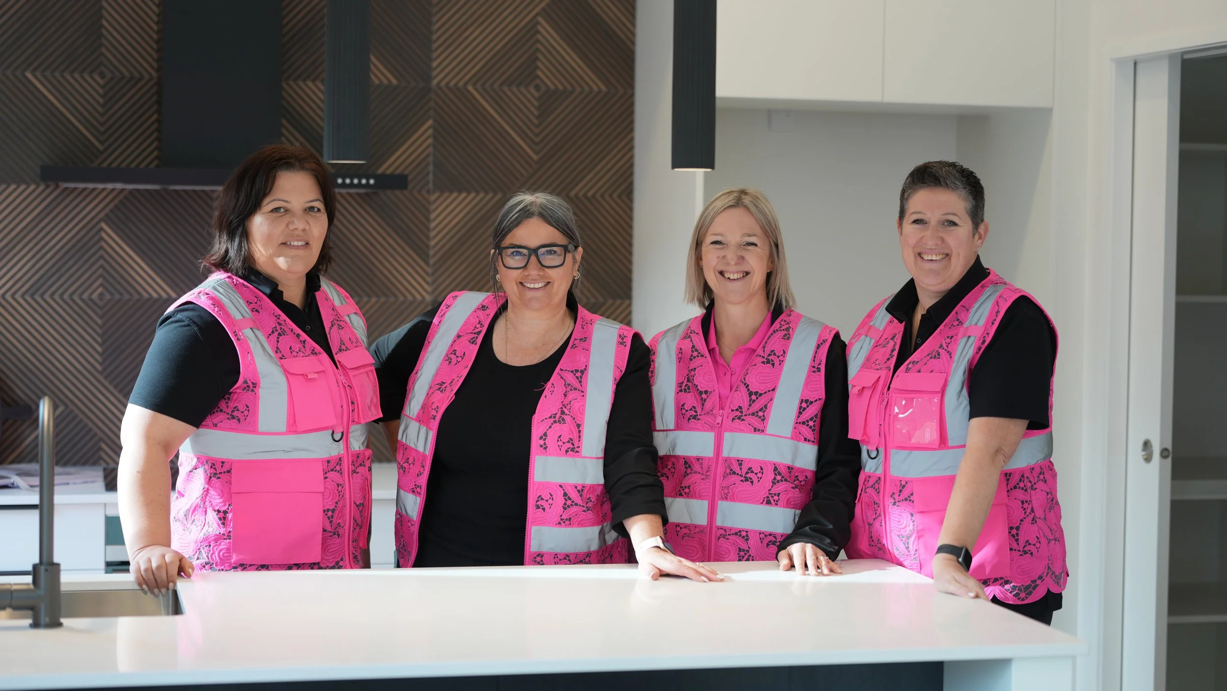 Four women smiling and wearing pink safety vests with reflective stripes standing around a white kitchen island.