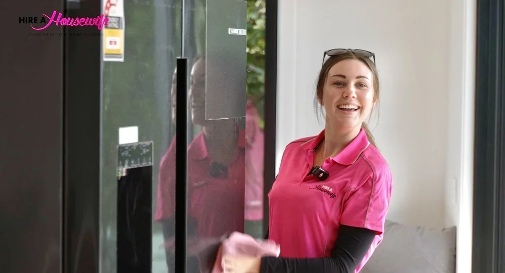 A woman in a pink uniform smiling while holding a tray, standing next to a refrigerator in a well-lit room.