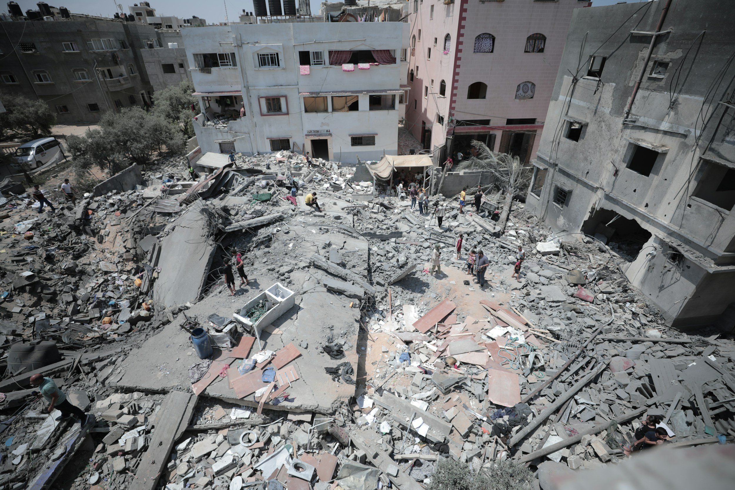Buildings and street heavily damaged with rubble and debris, indicating destruction from an earthquake or explosion; people are seen among the wreckage.