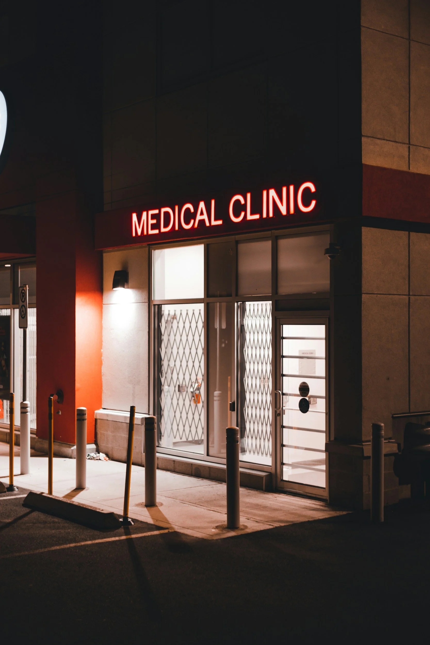Night view of a medical clinic entrance with a red illuminated sign reading 'Medical Clinic' above glass doors. Bollards and parking lot in the foreground.