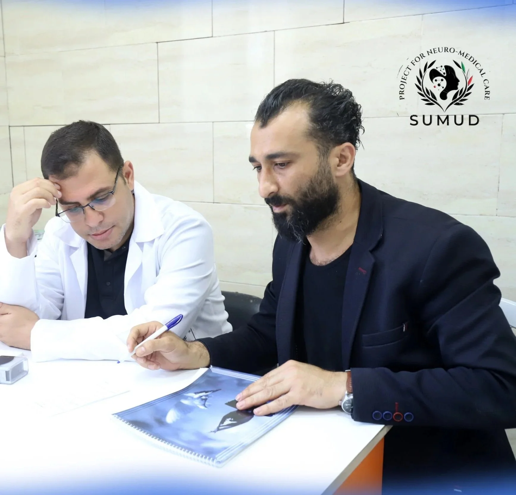 Dr. Ahmed Alkahlout sitting at a table reviewing documents, one in a white coat and the other in a black blazer, in a medical or professional setting with a logo for SUMUD Project for Neuro-Medical Care - SUMUD on the wall.