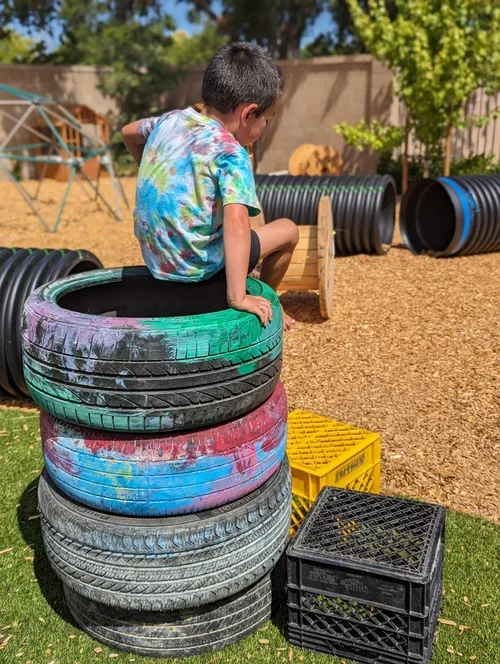 A child sitting inside a stack of colorful tires in a backyard playground area with black tubing, a yellow crate, and a black crate nearby, under clear blue skies.