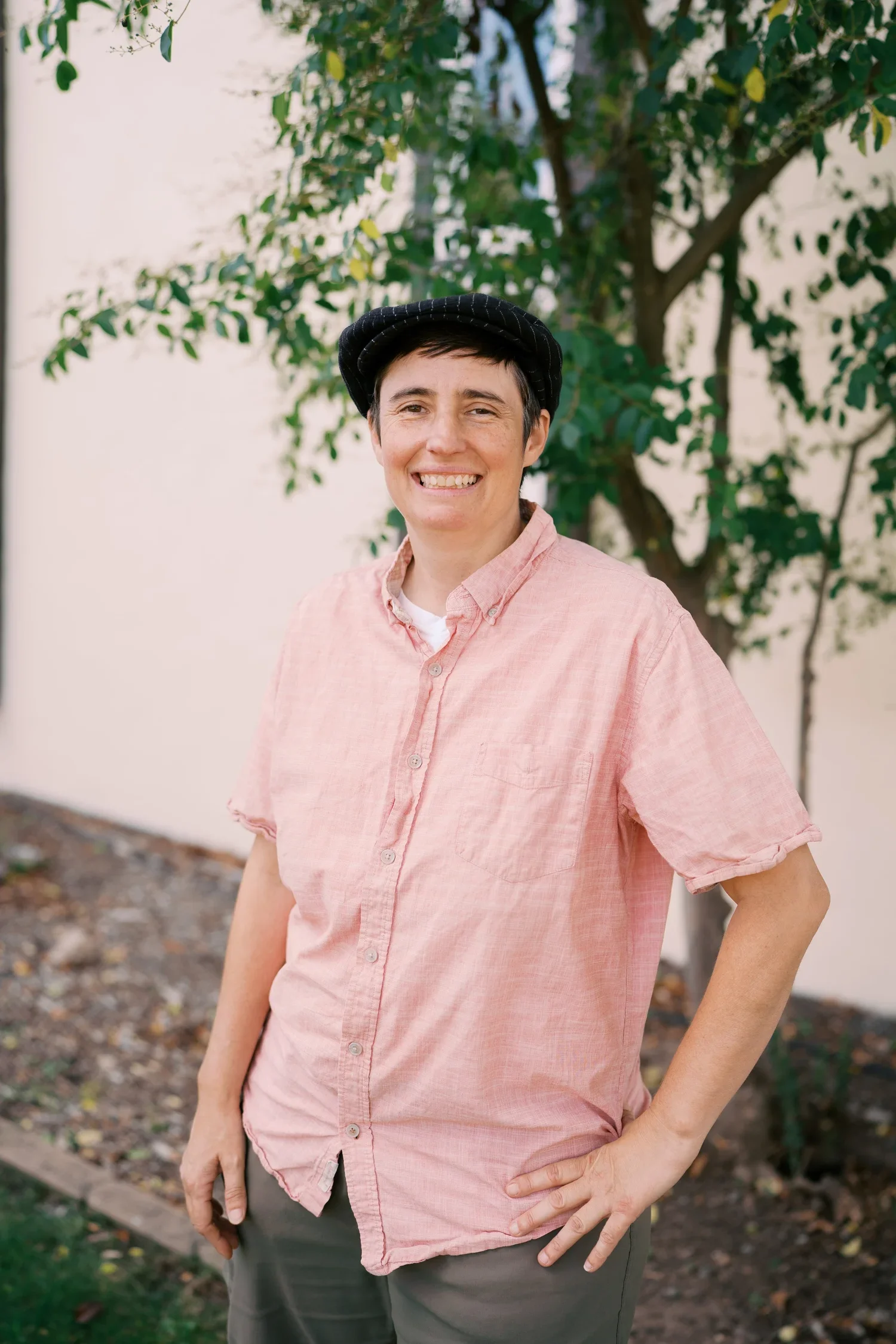 A person with short dark hair wearing a black cap, pink button-up shirt, and gray pants standing outdoors near a tree with green leaves.