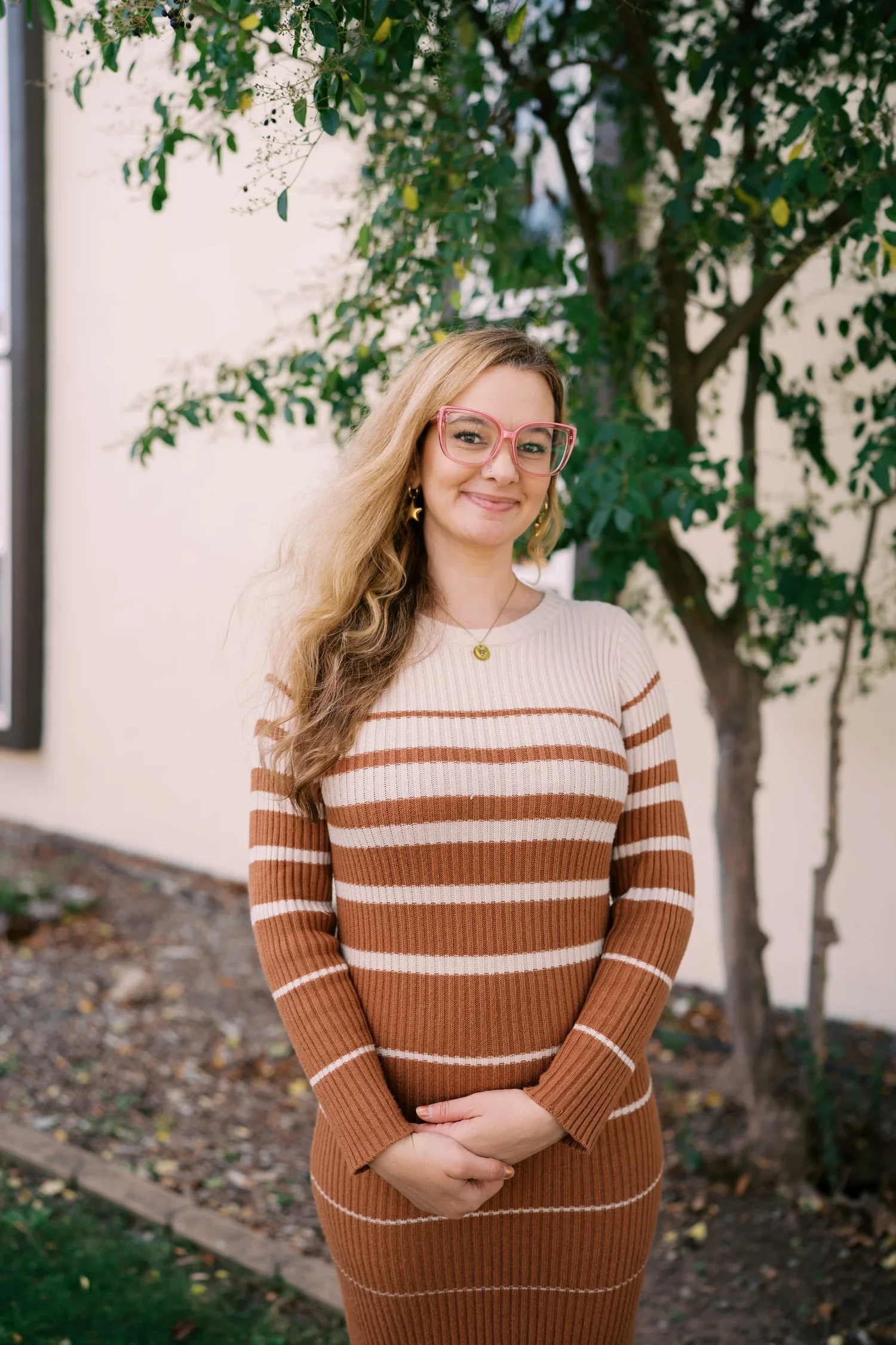 A woman with long wavy blonde hair wearing pink glasses, gold star-shaped earrings, a gold necklace, and a brown and beige striped sweater dress standing outdoors with a tree in the background.