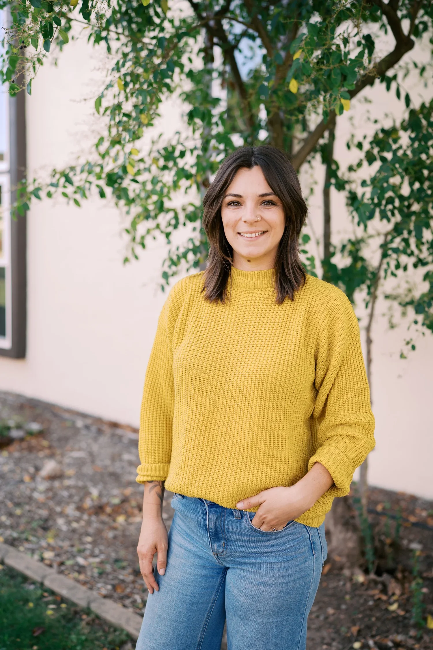 A woman standing outdoors near a tree, wearing a yellow sweater and blue jeans, smiling at the camera.