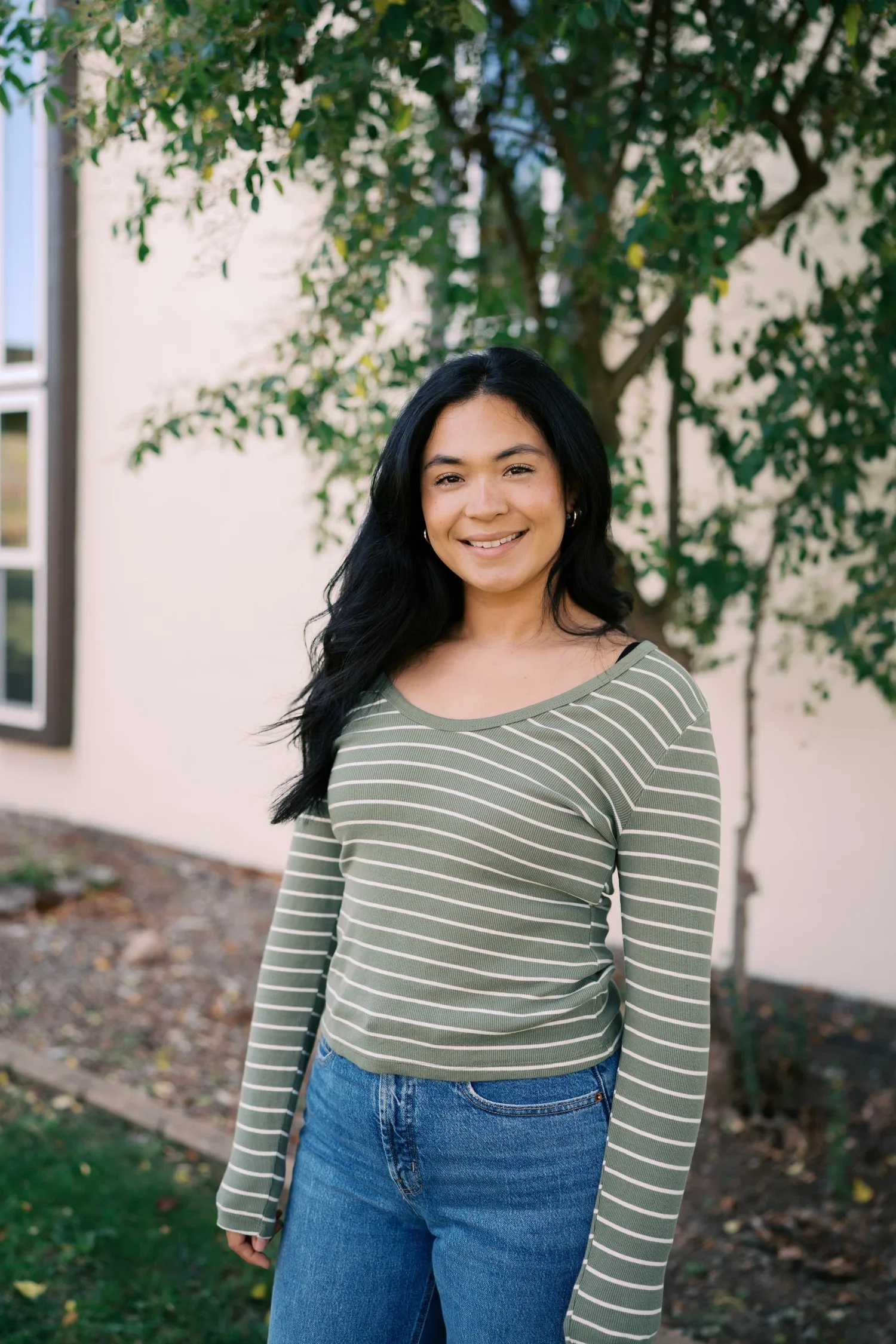 A young woman with long black hair, wearing a green and white striped long-sleeve shirt and blue jeans, standing outdoors in front of a tree with green leaves and a beige wall.