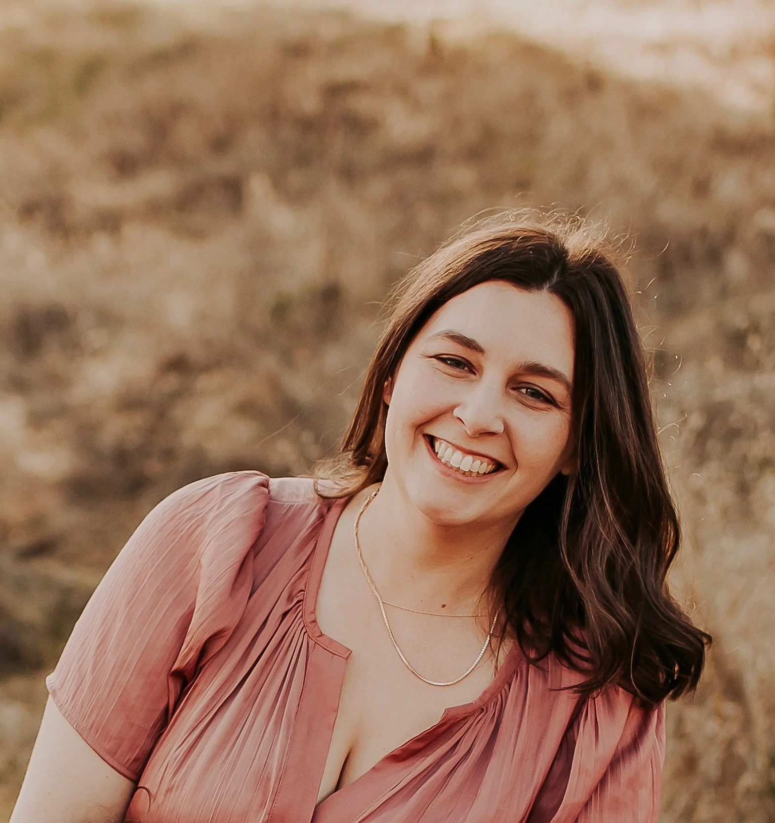 A young woman with brown hair smiling outdoors, wearing a pink dress and a necklace, with a blurred natural background.
