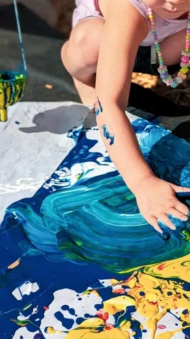 Child with a necklace playing with blue and yellow paint on paper outdoors.