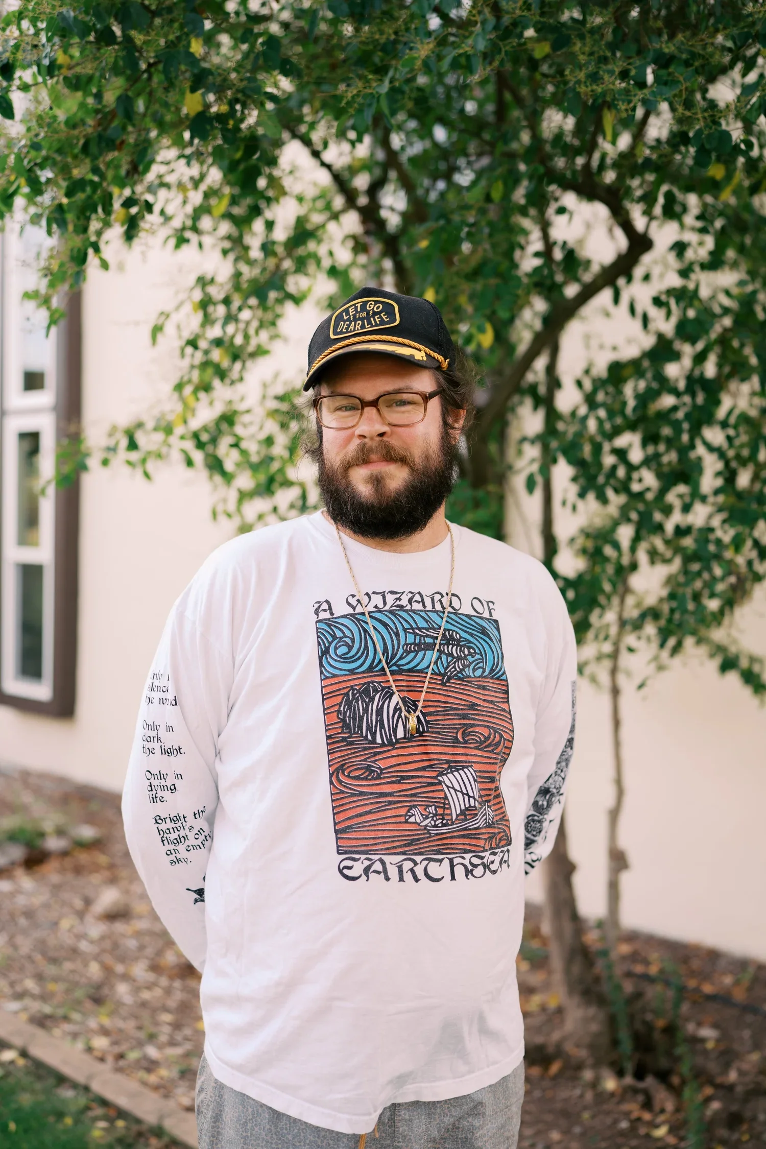 A man with glasses and a beard wearing a black cap with yellow accents and text, a white long-sleeve graphic shirt featuring stylized ocean waves and a boat, standing outdoors in front of a tree and building, with fallen leaves on the ground.