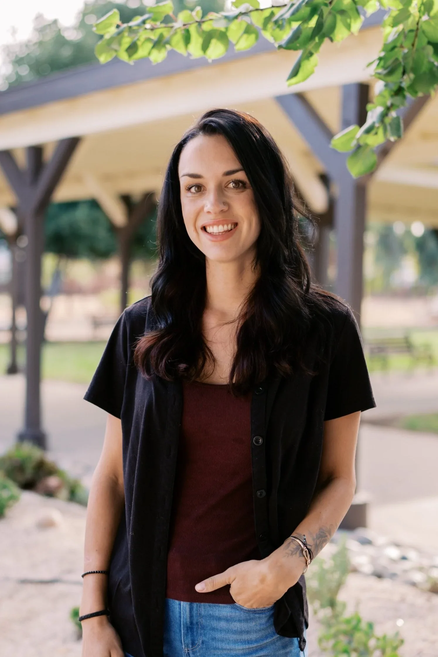 A woman with long dark hair, wearing a dark shirt over a maroon top and blue jeans, standing outdoors under a covered structure with greenery around, smiling at the camera.