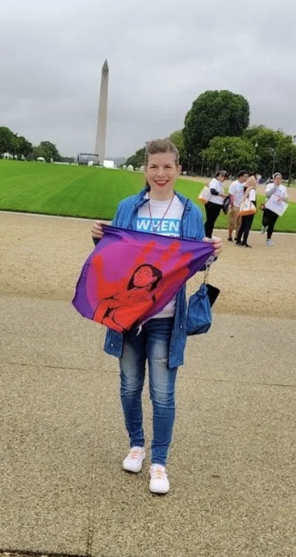A young woman with short hair, wearing a blue jacket and jeans, stands on a paved pathway with the Washington Monument visible in the background. She is smiling and holding a colorful flag featuring an illustration of a woman with long hair and red clothing. There are a few people walking in the background on a cloudy day.