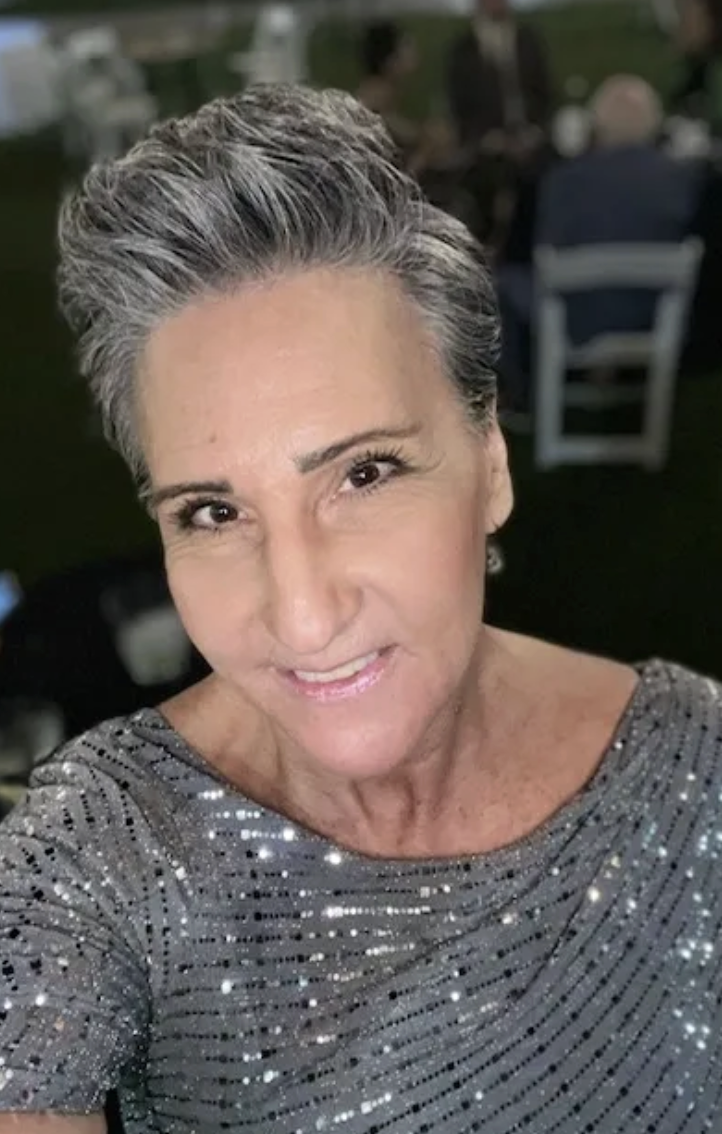 A close-up photo of a woman with short gray hair wearing a silver, sequined top. She is smiling and making a slight wink.