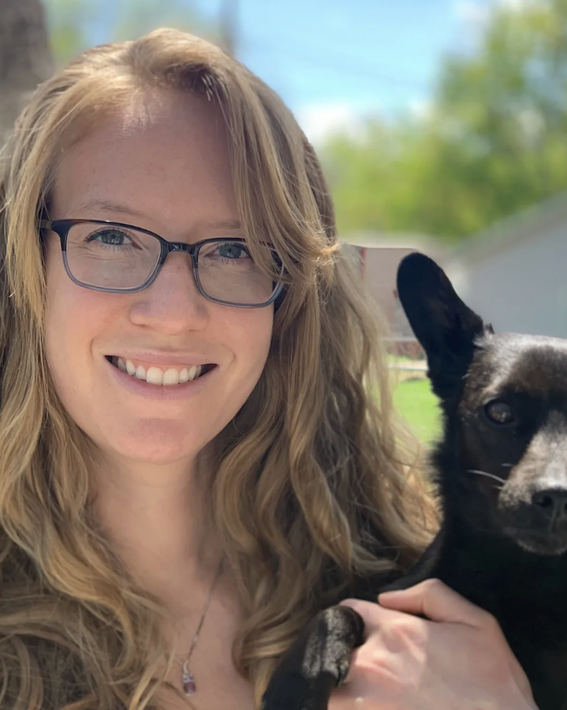 A woman with glasses smiling and holding a black dog.