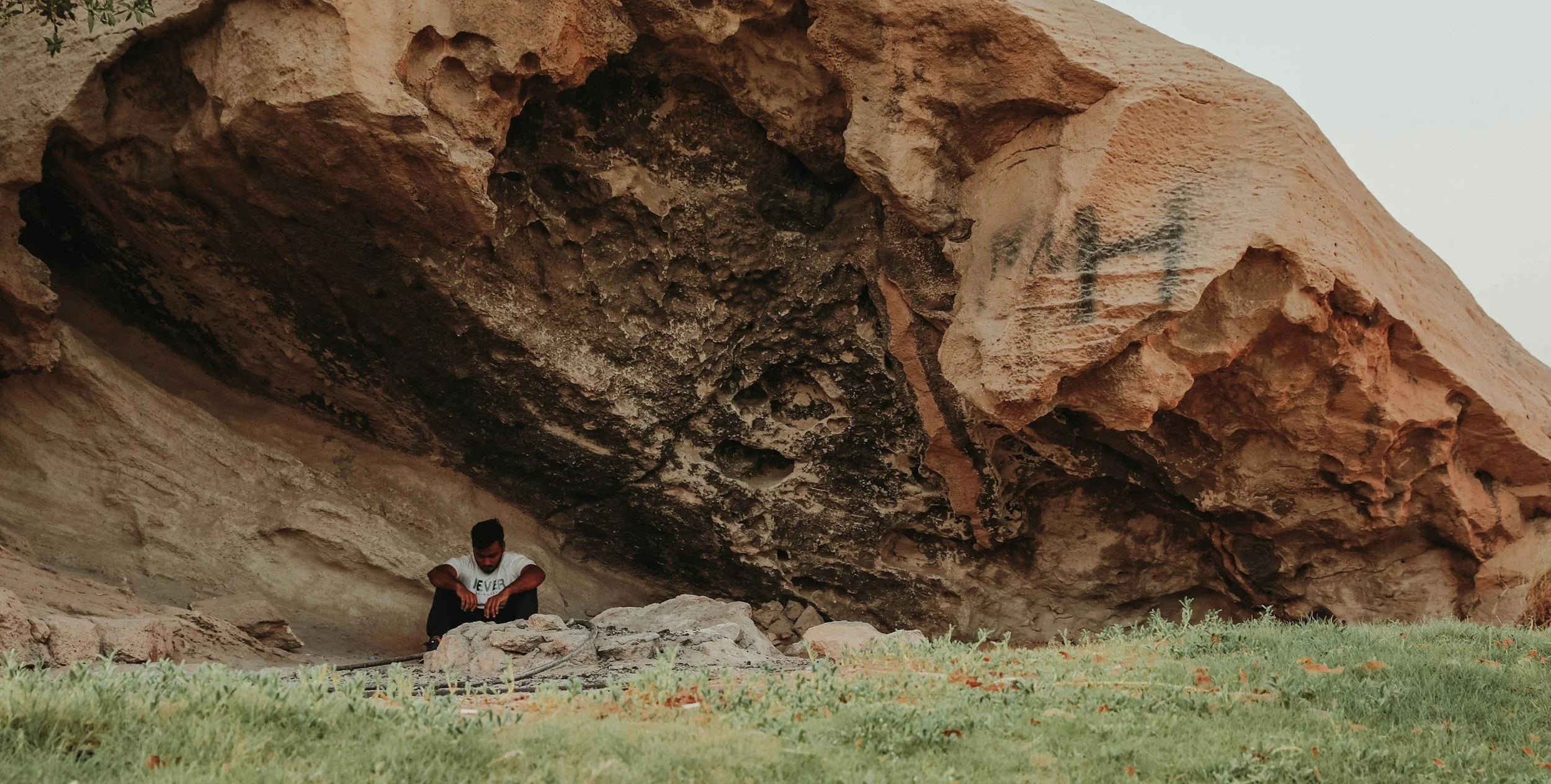 A person sitting under a large, overhanging rock formation with a textured underside in a natural outdoor setting. The person appears to be resting or contemplating, with grass and small plants visible at the base.