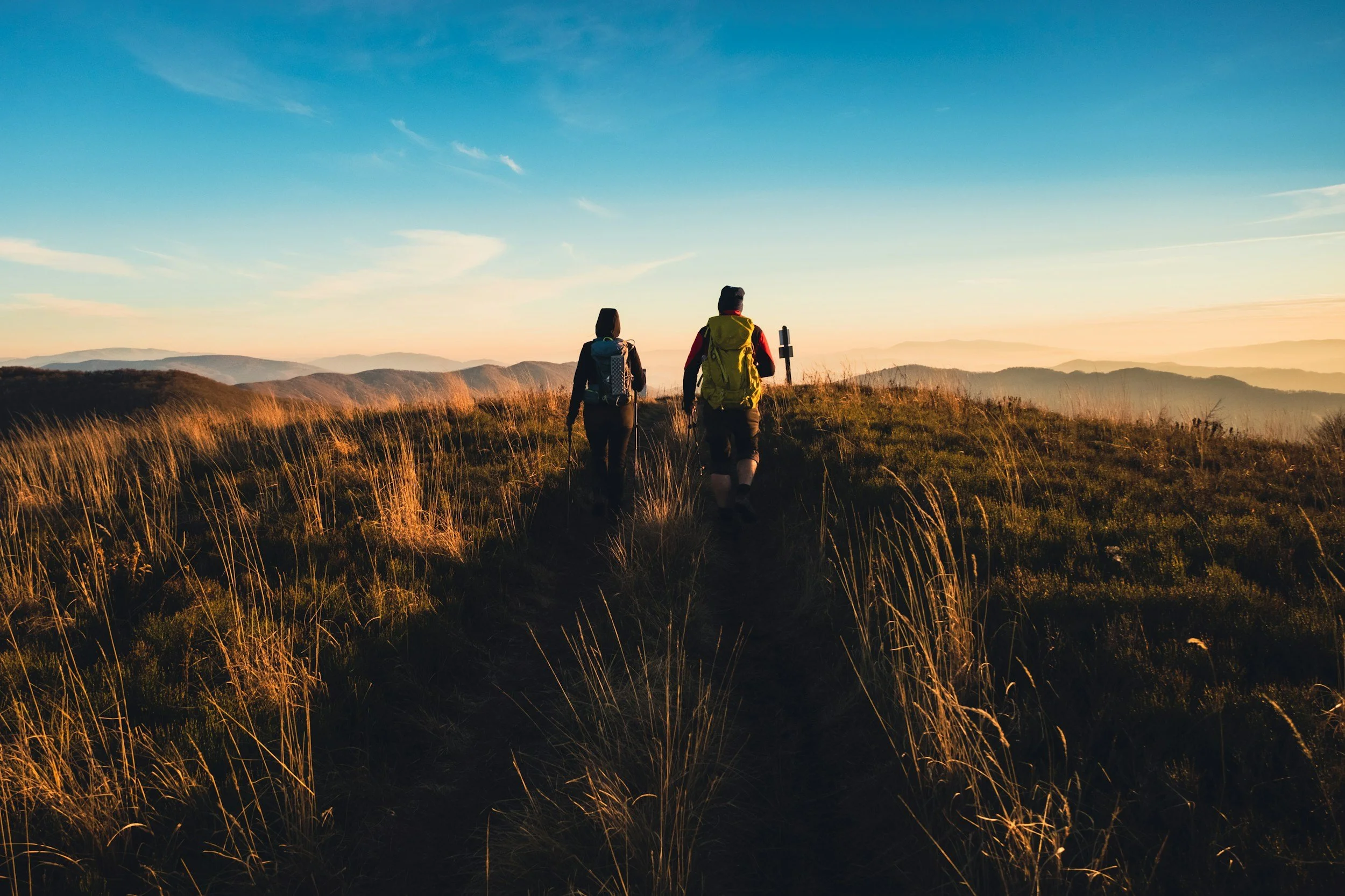 Two hikers walking on a trail in the mountains during sunset, with rolling hills and a clear sky in the background.