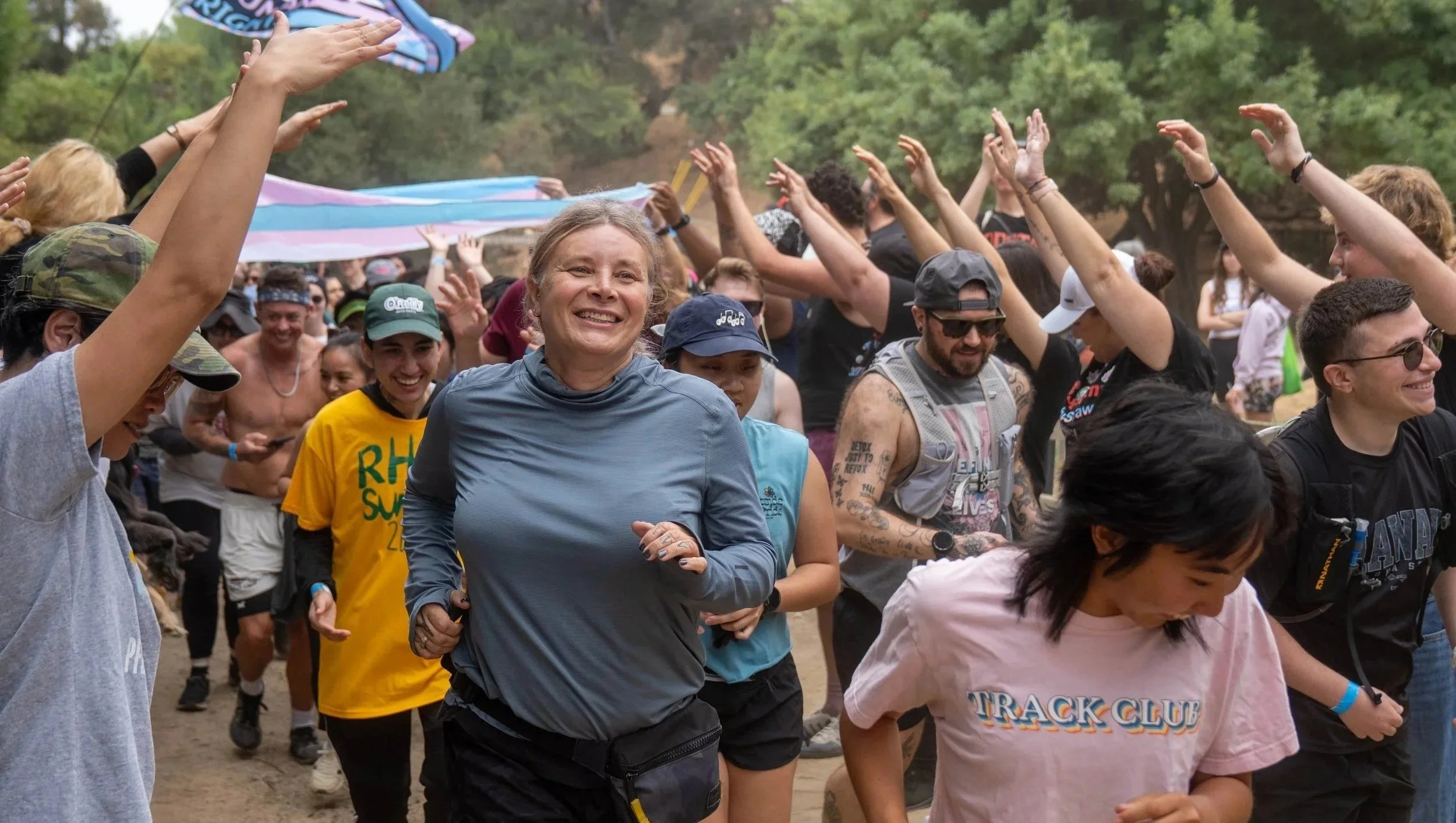 A line of people run happily through outstretched arms and Trans Pride flags at the Trans Pride 5K