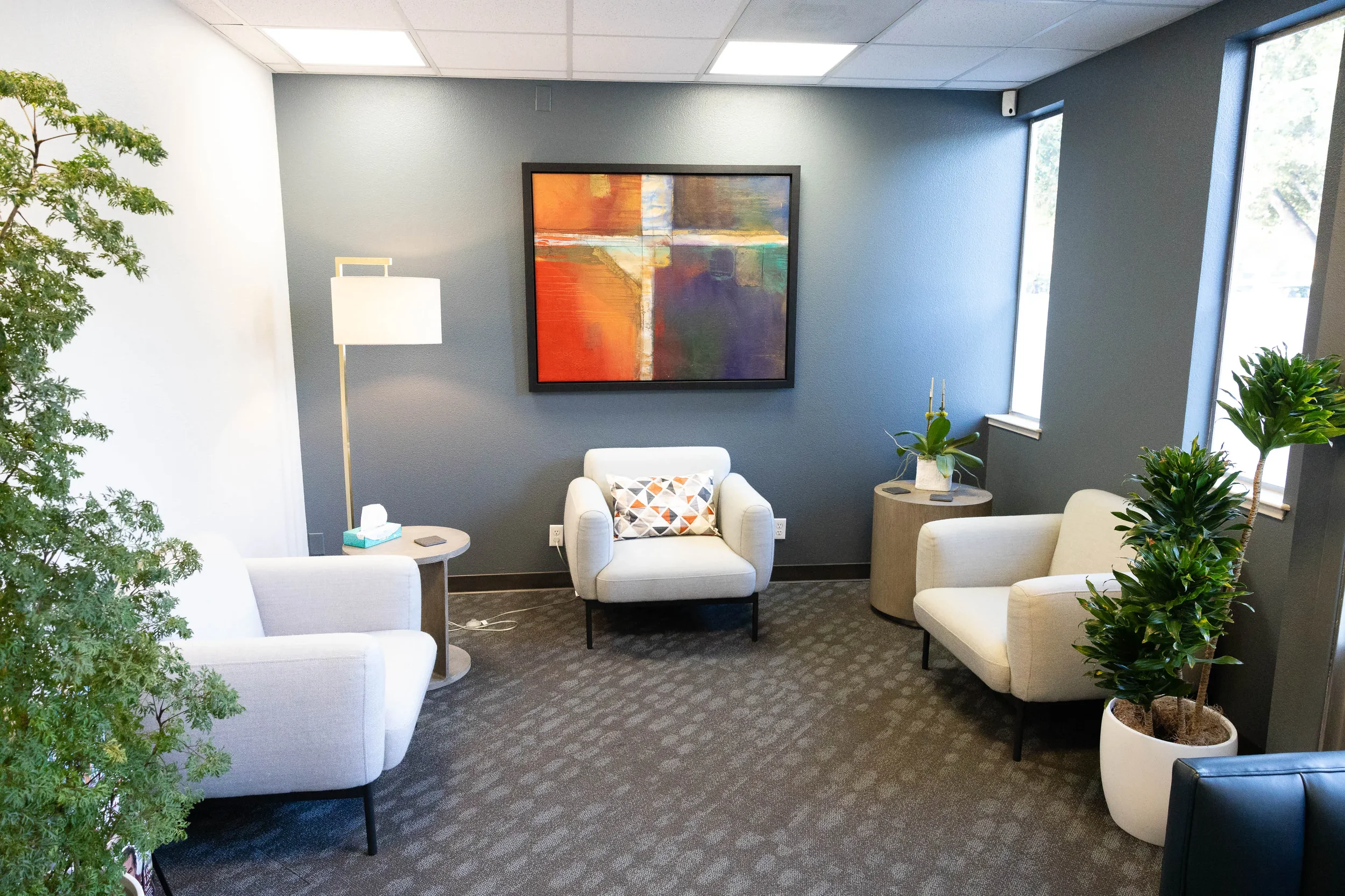 A cozy waiting area with white armchairs, a small side table, artwork, and indoor plants
