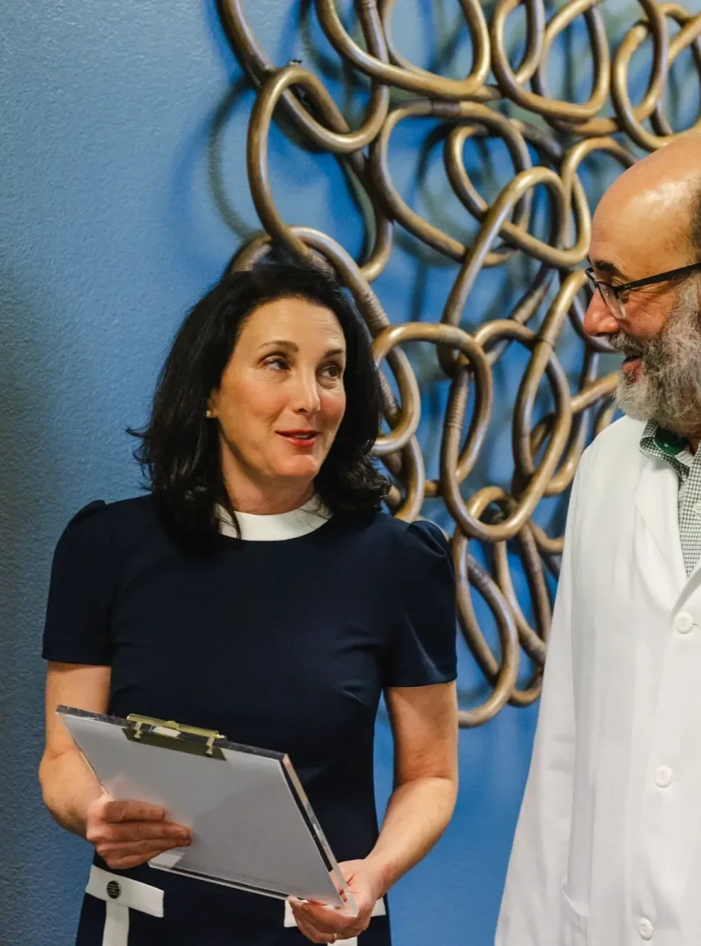 Concierge doctor Bonnie Rosenberg speaking with a man in front of blue wall with a gold metal art piece.