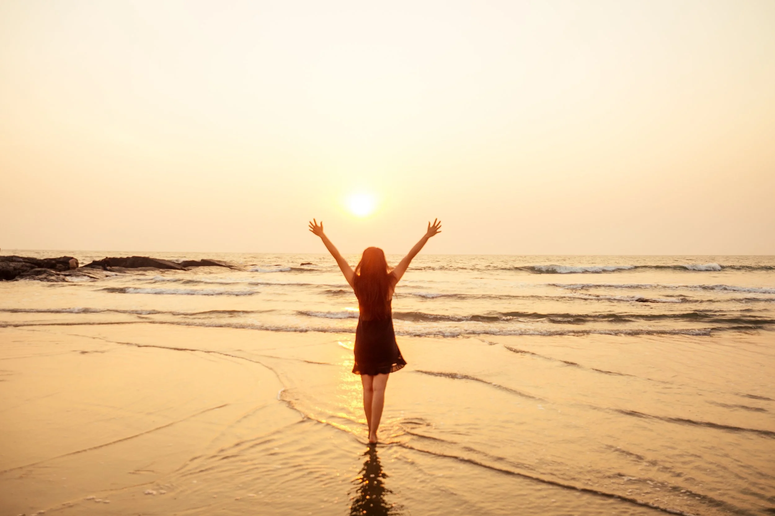 A woman standing on the beach with her arms raised towards the sunset over the ocean.