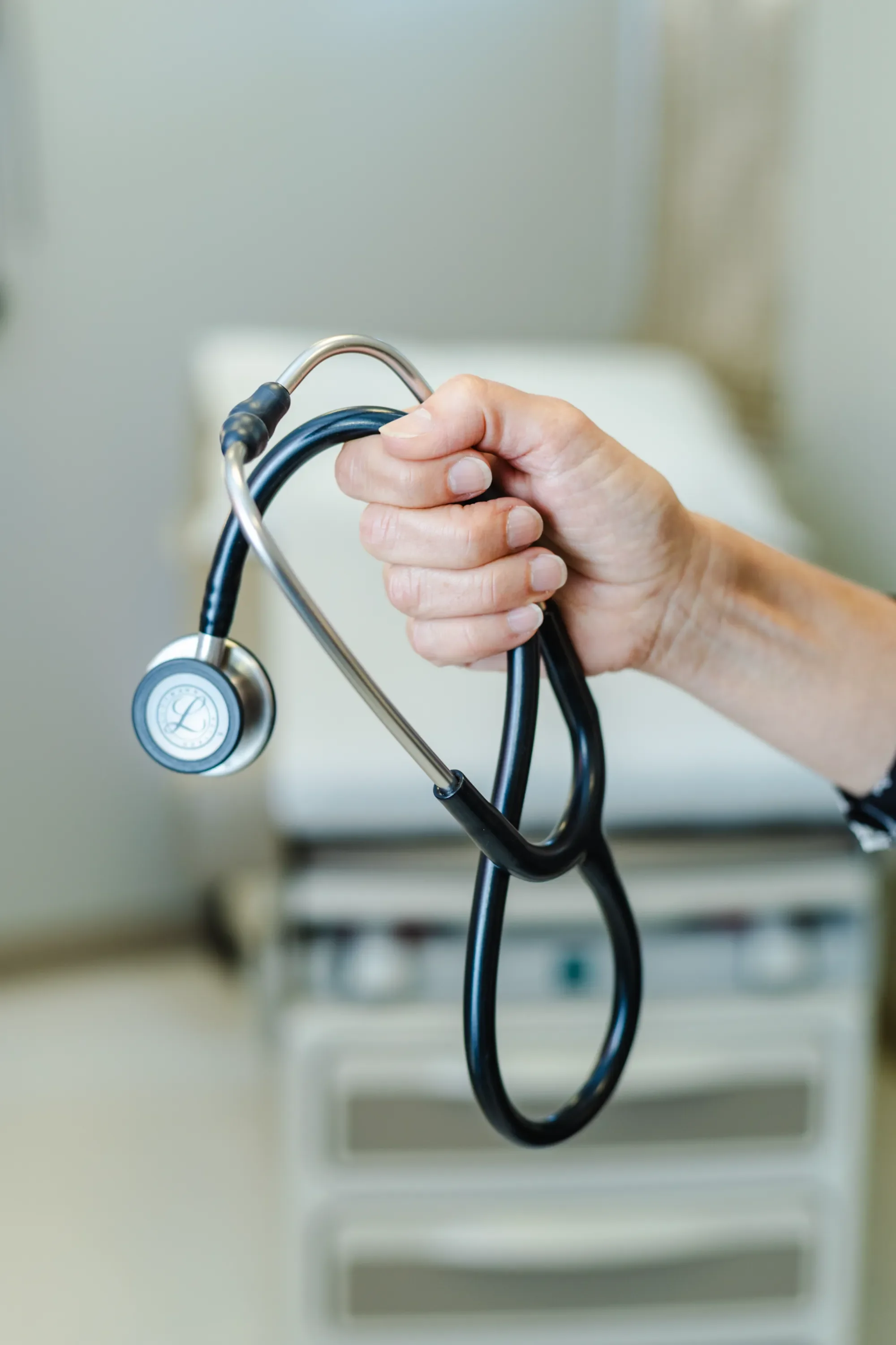 A hand holding a stethoscope' with a blurred background of a medical office.