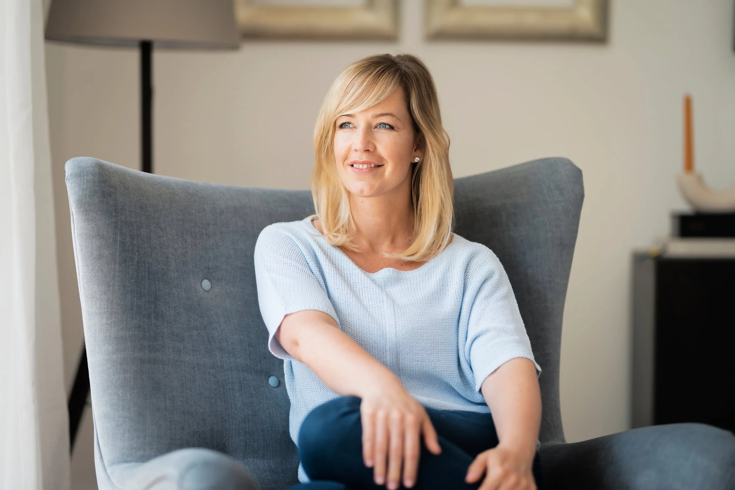 A woman with blonde hair in a gray armchair, smiling and wearing a light blue top.
