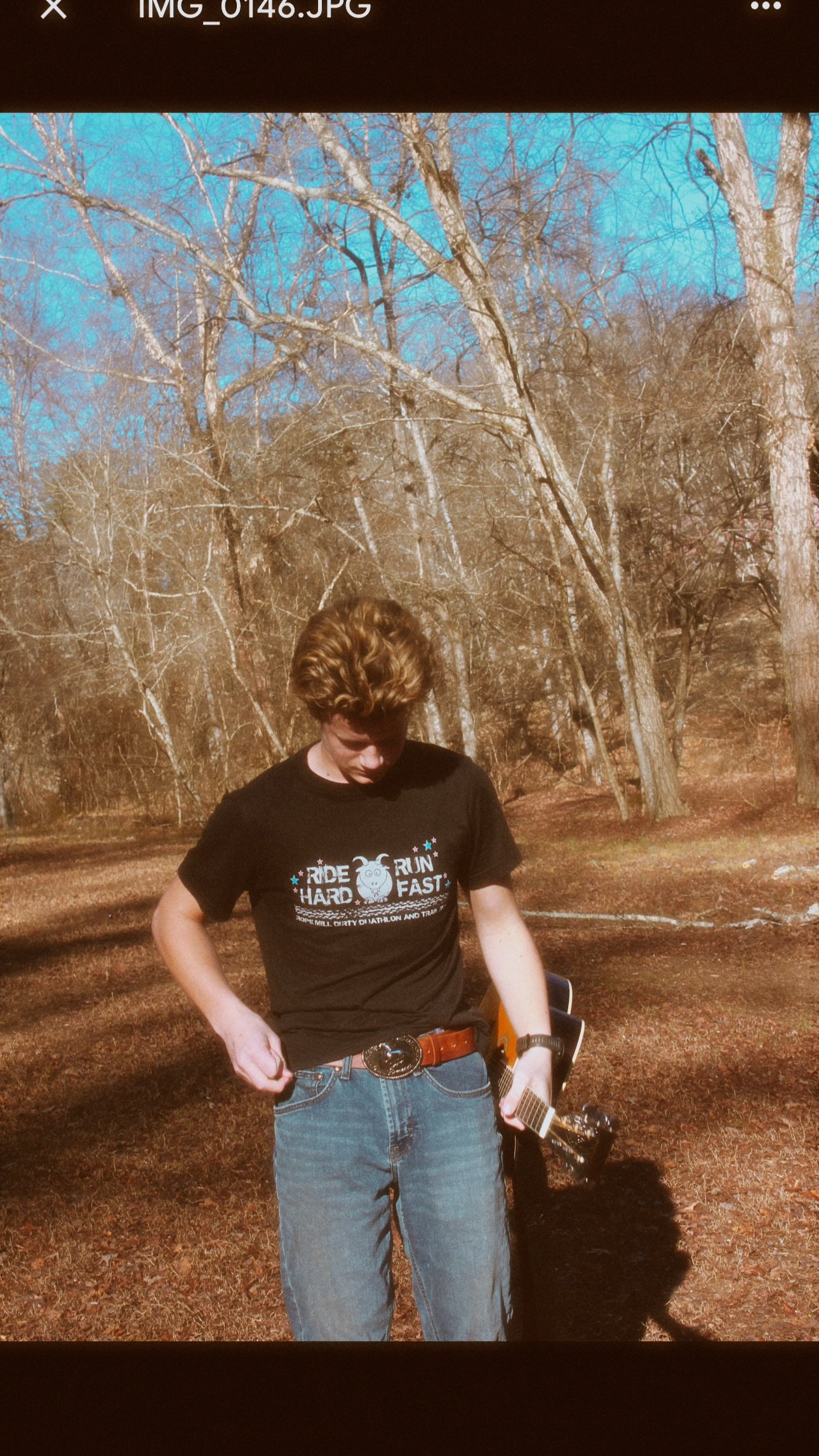 Young man with curly hair standing outdoors in a wooded area, holding a guitar, wearing a black T-shirt and jeans.