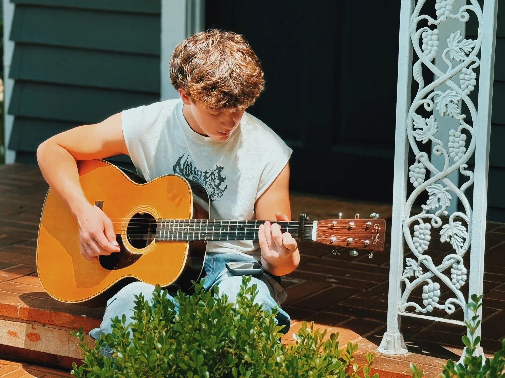 Young man with curly hair playing an acoustic guitar on a porch, sitting on brick steps surrounded by green plants, with a white decorative iron railing to his right.